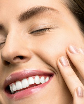 A close-up portrait of a joyful woman with closed eyes, pink lipstick, and her hand touching her cheek.